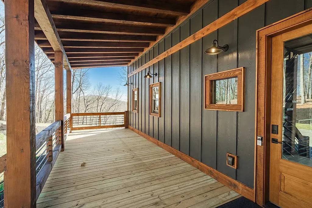 A covered wooden porch with a dark vertical-planked wall, a glass door, and a view of a treed landscape.