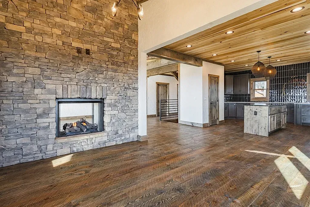 A living room with a large stone fireplace, exposed wooden beam ceiling, and a view into a rustic kitchen.