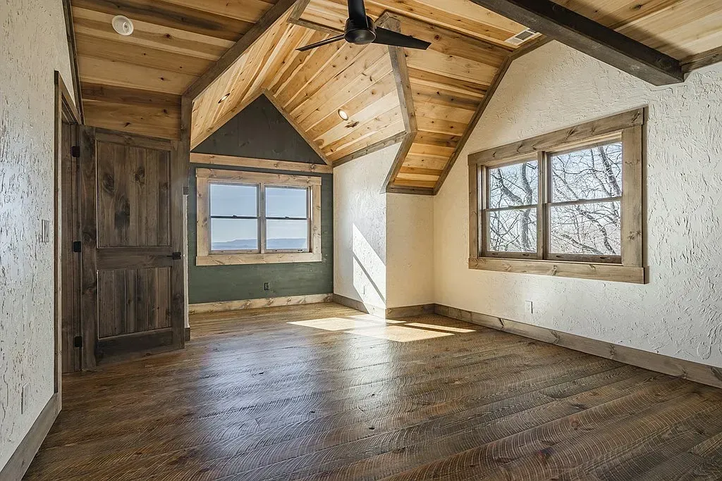 A rustic bedroom with wood-paneled vaulted ceilings, light textured walls, wooden floors, and two large windows.