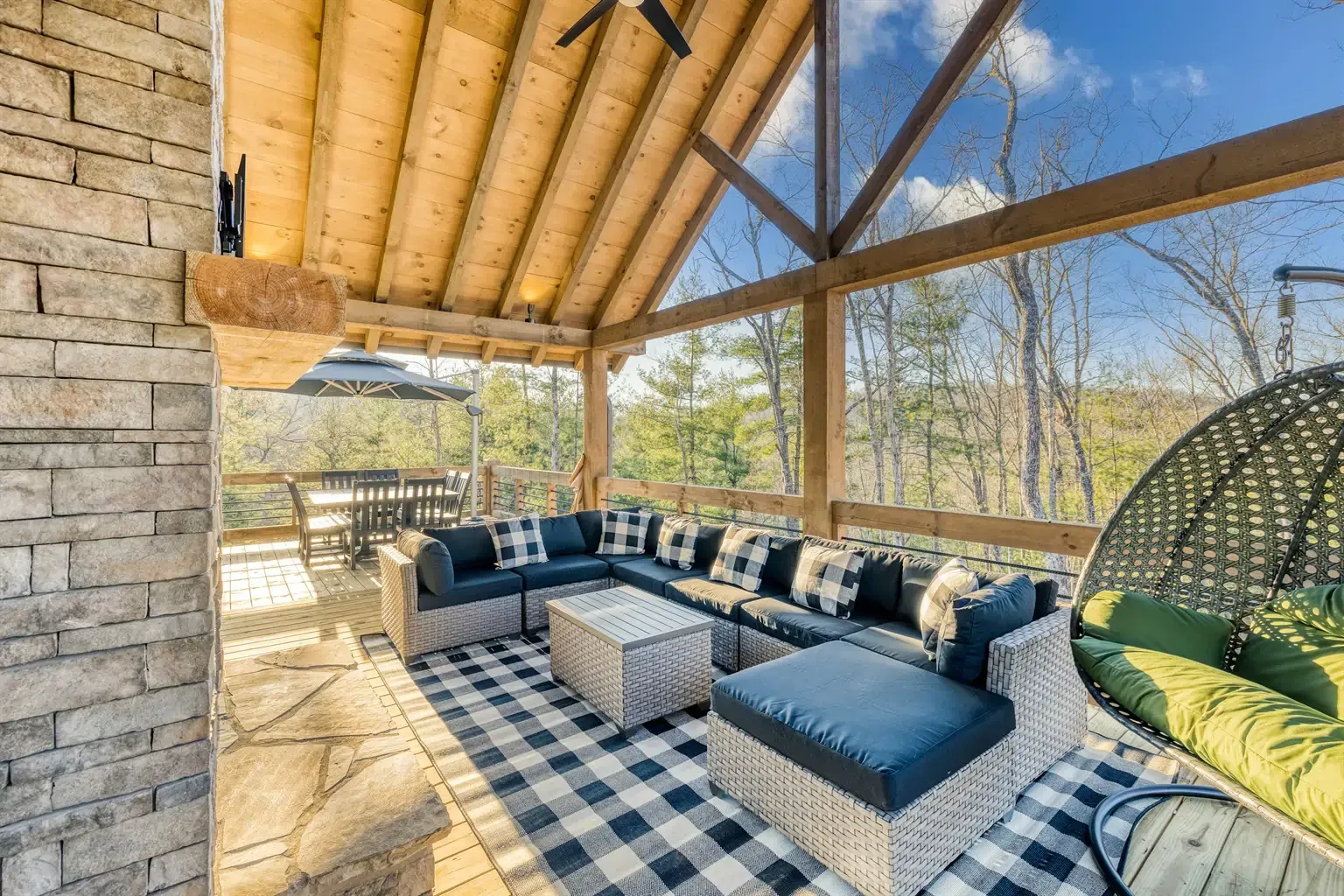 A screened-in porch with a stone fireplace, wicker sectional sofa, black and white rug, and views of a forested hillside.