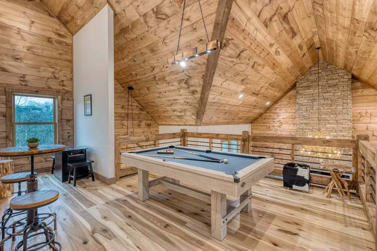 A rustic loft space features a pool table, wooden walls, a stone fireplace, and a small seating area with bar stools.