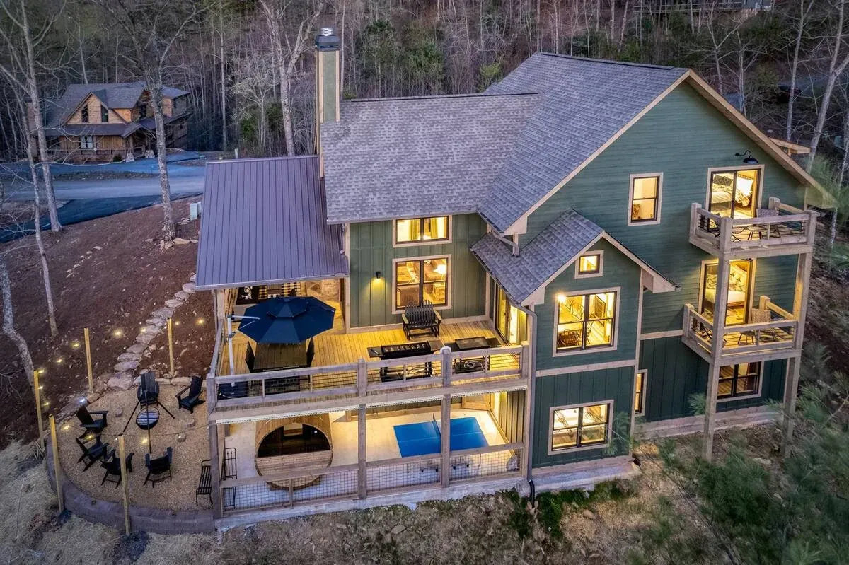 An aerial view of a multi-story green cabin in a forest, featuring a deck with patio furniture, a fire pit, and a pool.