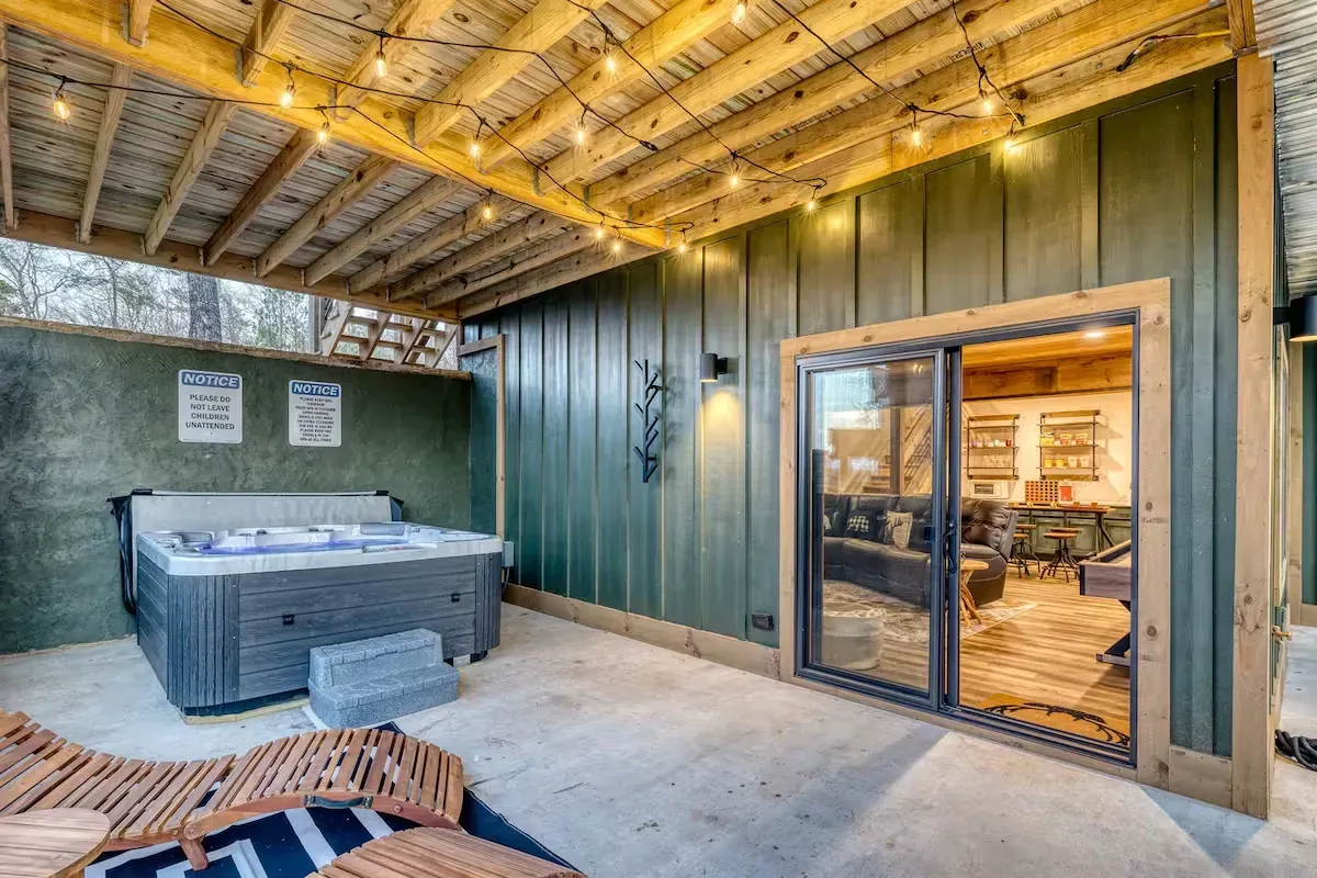 A covered outdoor patio featuring a hot tub, a wooden lounge chair, string lights, and a sliding glass door.