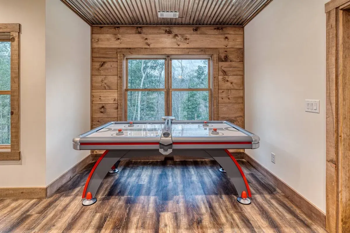An air hockey table stands in a rustic room with wood-paneled walls, a corrugated metal ceiling, and a view of trees.