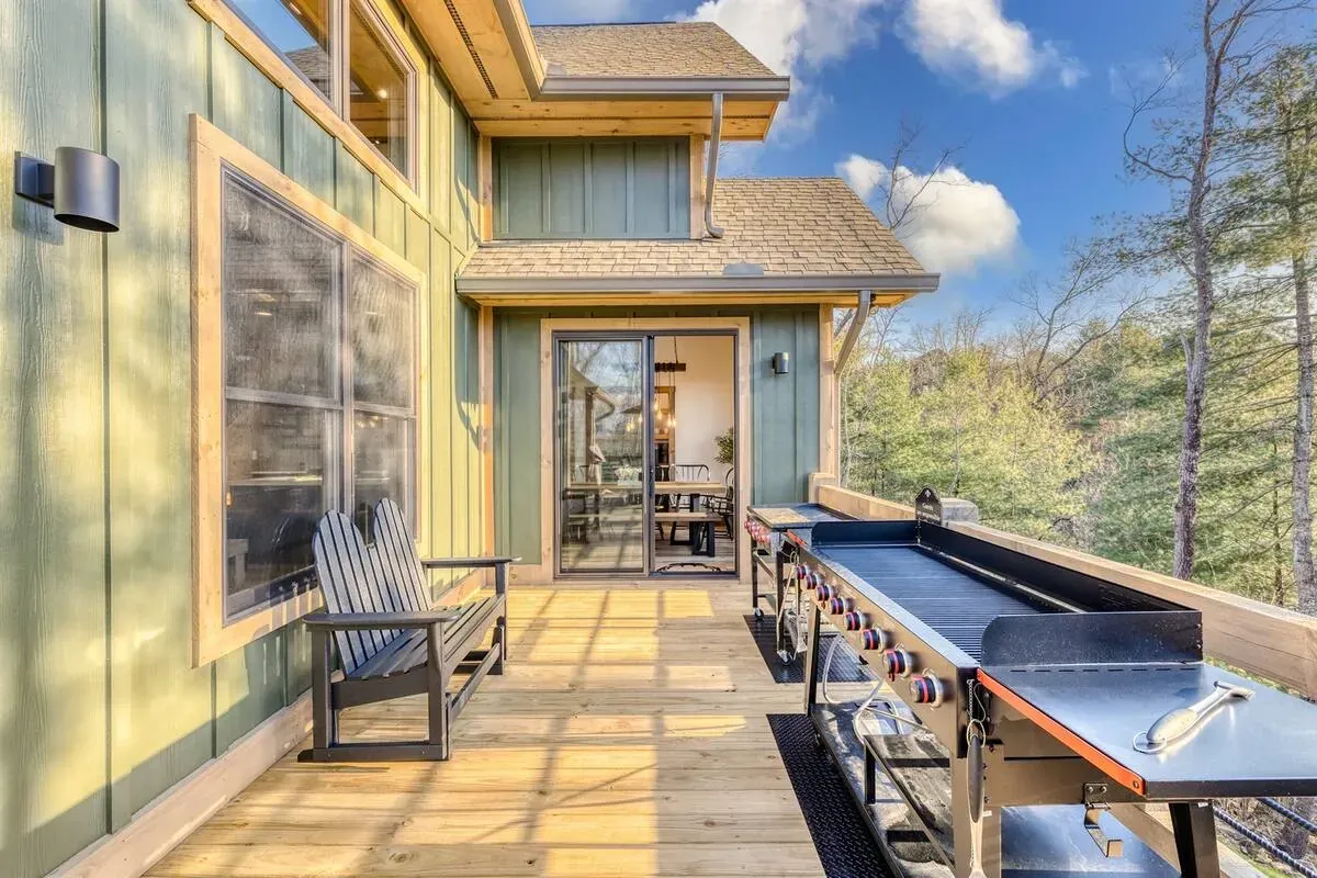 A wooden deck attached to a sage green house featuring a large outdoor griddle and a bench, overlooking a forest.