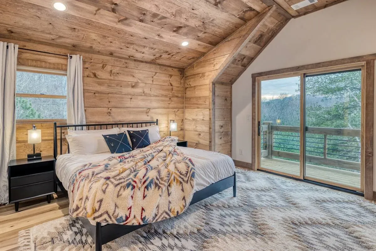 A rustic bedroom with wooden walls and ceiling, a queen bed with a patterned throw, and a large sliding glass door.