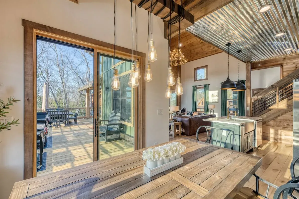 A rustic dining room featuring a wooden table, large glass sliding doors to a deck, and hanging lights under a metal roof.