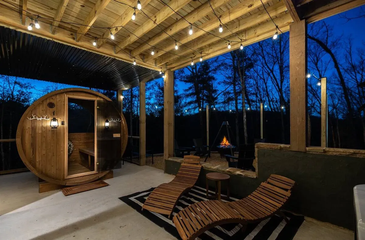 A barrel sauna on a lighted patio at dusk, featuring two wooden lounge chairs on a black-and-white rug.