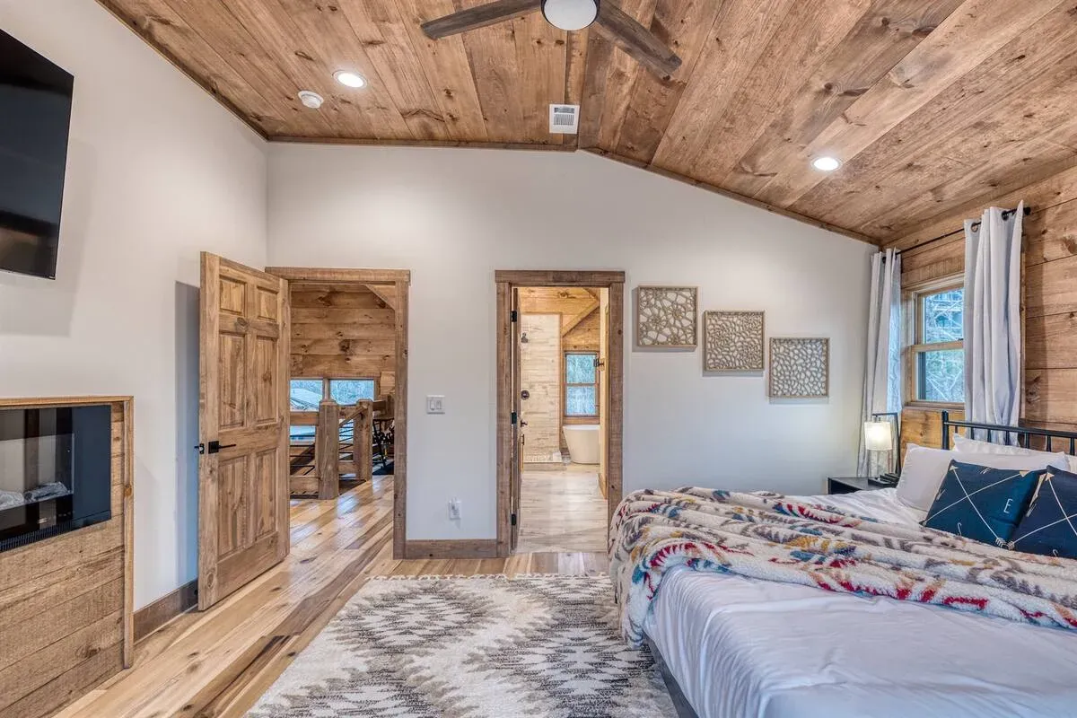 A bedroom with a wooden plank ceiling, a bed with a patterned quilt, a rug, and two doorways leading into other rooms.