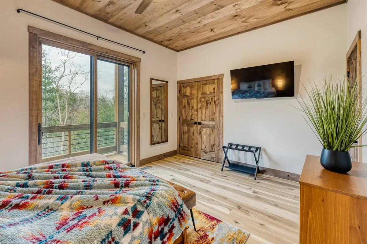 A bedroom with a rustic wood ceiling, sliding glass doors to a balcony, a patterned blanket on the bed, and a wall TV.