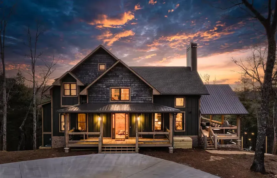 A dark-sided two-story cabin with a wrap-around porch at dusk, set against a colorful, dramatic sunset sky.