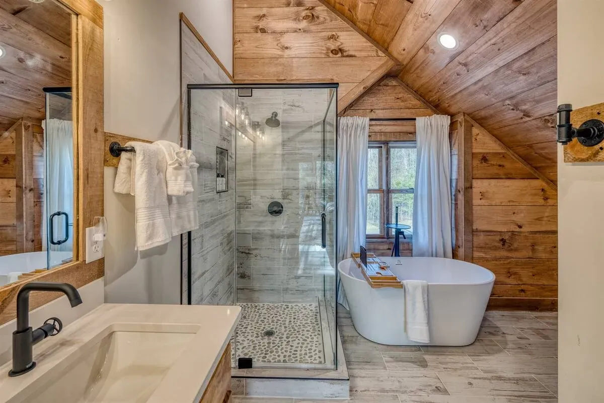 A rustic bathroom with a white freestanding tub, a glass-enclosed shower, a vanity, and wood-paneled walls and ceiling.