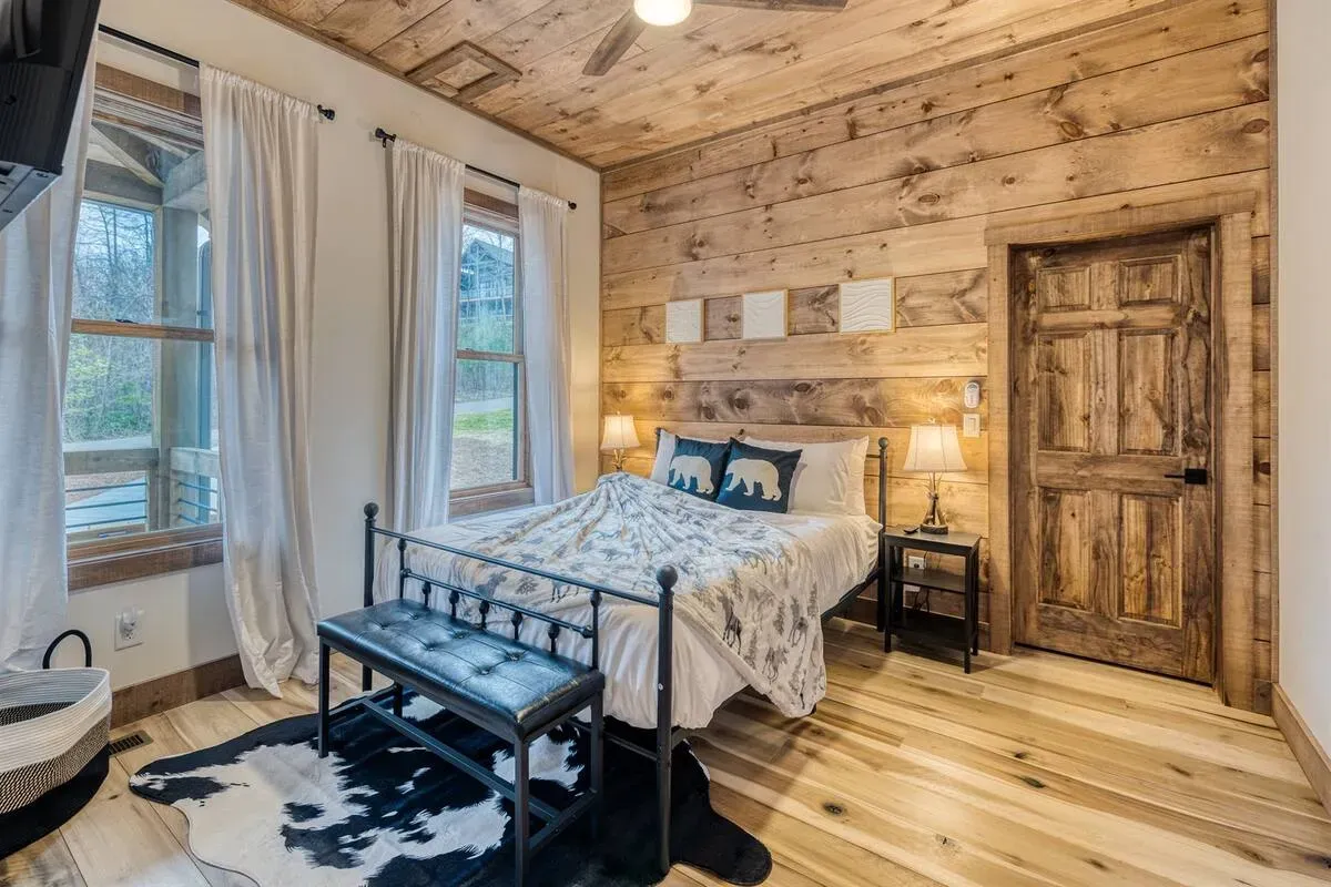 A rustic bedroom with a metal-framed bed, wood-paneled walls, a ceiling fan, a cowhide rug, and a wooden door.