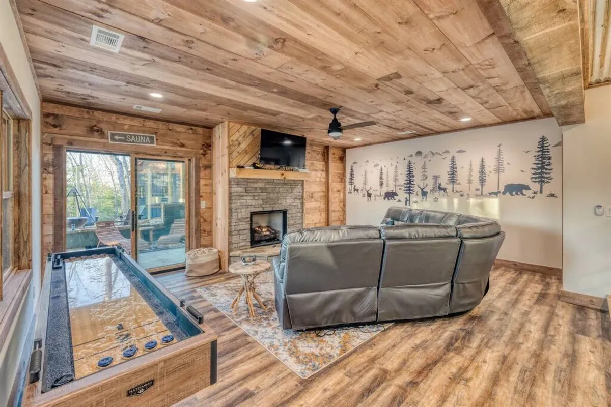 A rustic living room with wood paneled walls, a fireplace, a grey sectional sofa, and a shuffleboard table.