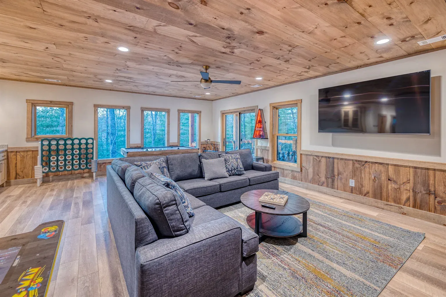 A rustic living room with a large gray sectional sofa, a round coffee table, a wooden ceiling, and a wall-mounted TV.