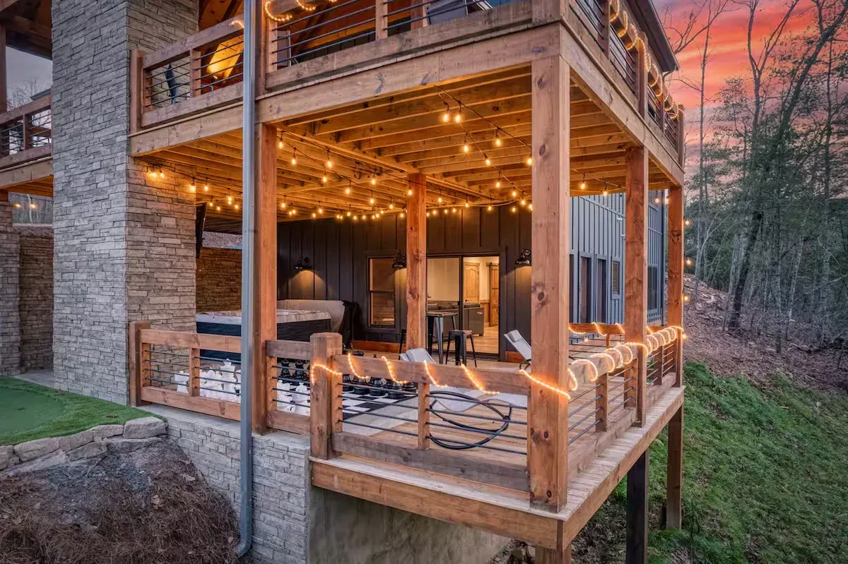 A rustic wooden deck at dusk with string lights, outdoor furniture, and a stone wall, overlooking a wooded hillside.