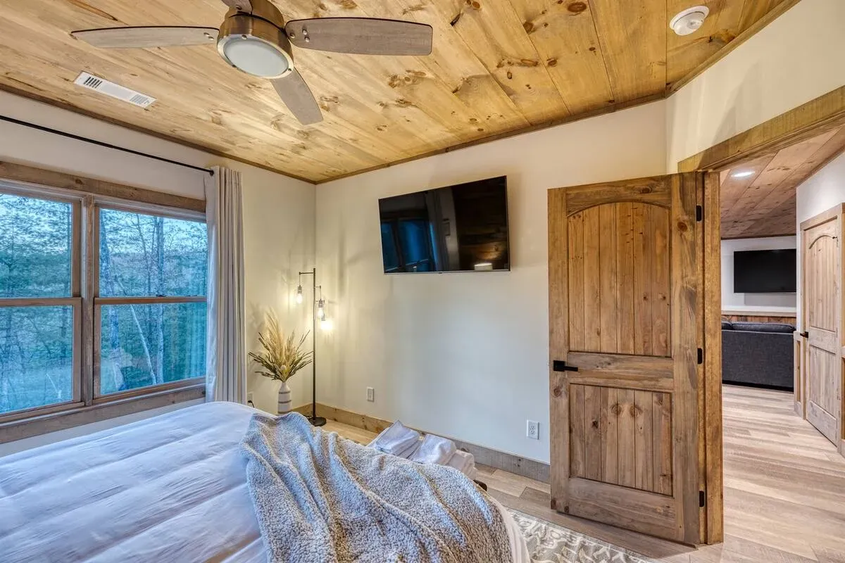 A cozy rustic bedroom with wood-paneled ceiling, a white bed, television on the wall, and an open wooden door.