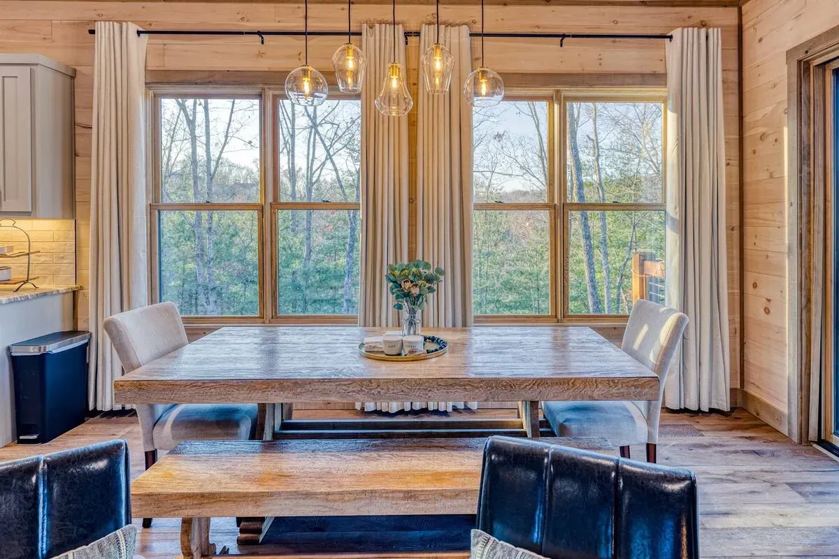 A rustic dining room with a wooden table, bench, and chairs, set before a large window with white curtains and lights.
