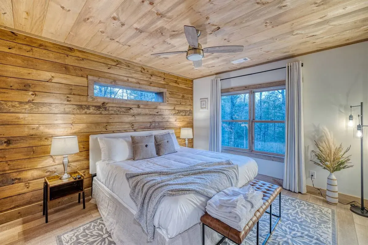 A cozy bedroom with a log-style wall, a white bed with a textured throw, a ceiling fan, and a floor lamp by a window.