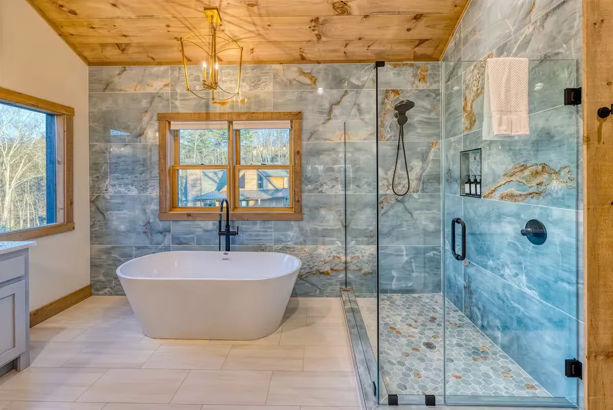 A modern bathroom featuring a white freestanding tub, a glass-enclosed shower with pebble tile floor, and blue tiled walls.
