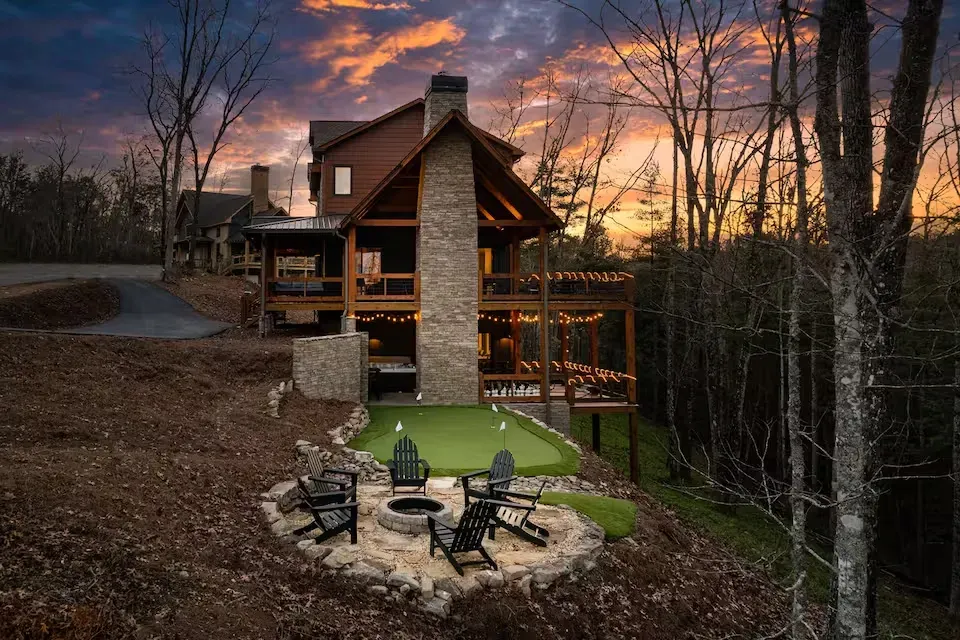 A multi-story wooden cabin at sunset with a stone chimney, backyard putting green, and a fire pit with outdoor chairs.