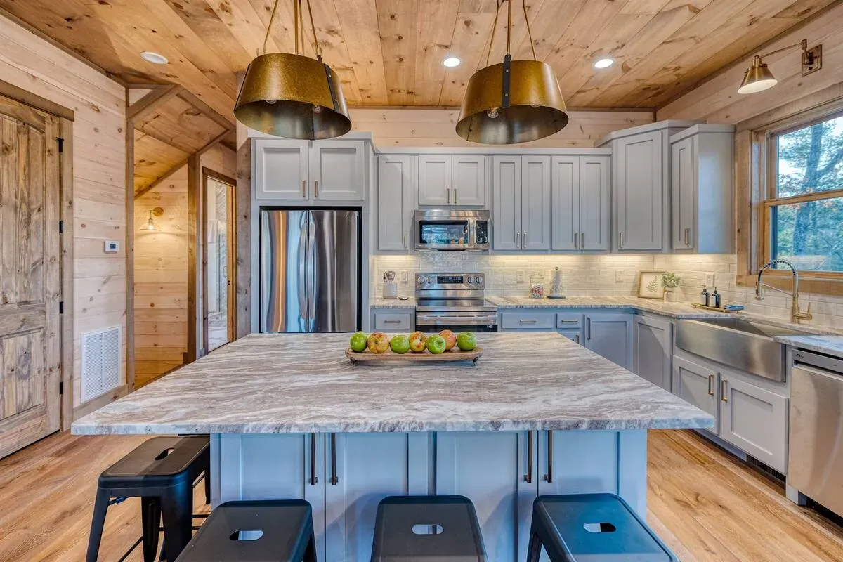 A bright rustic kitchen featuring light wood paneling, gray cabinetry, a large marble island, and dual pendant lights.