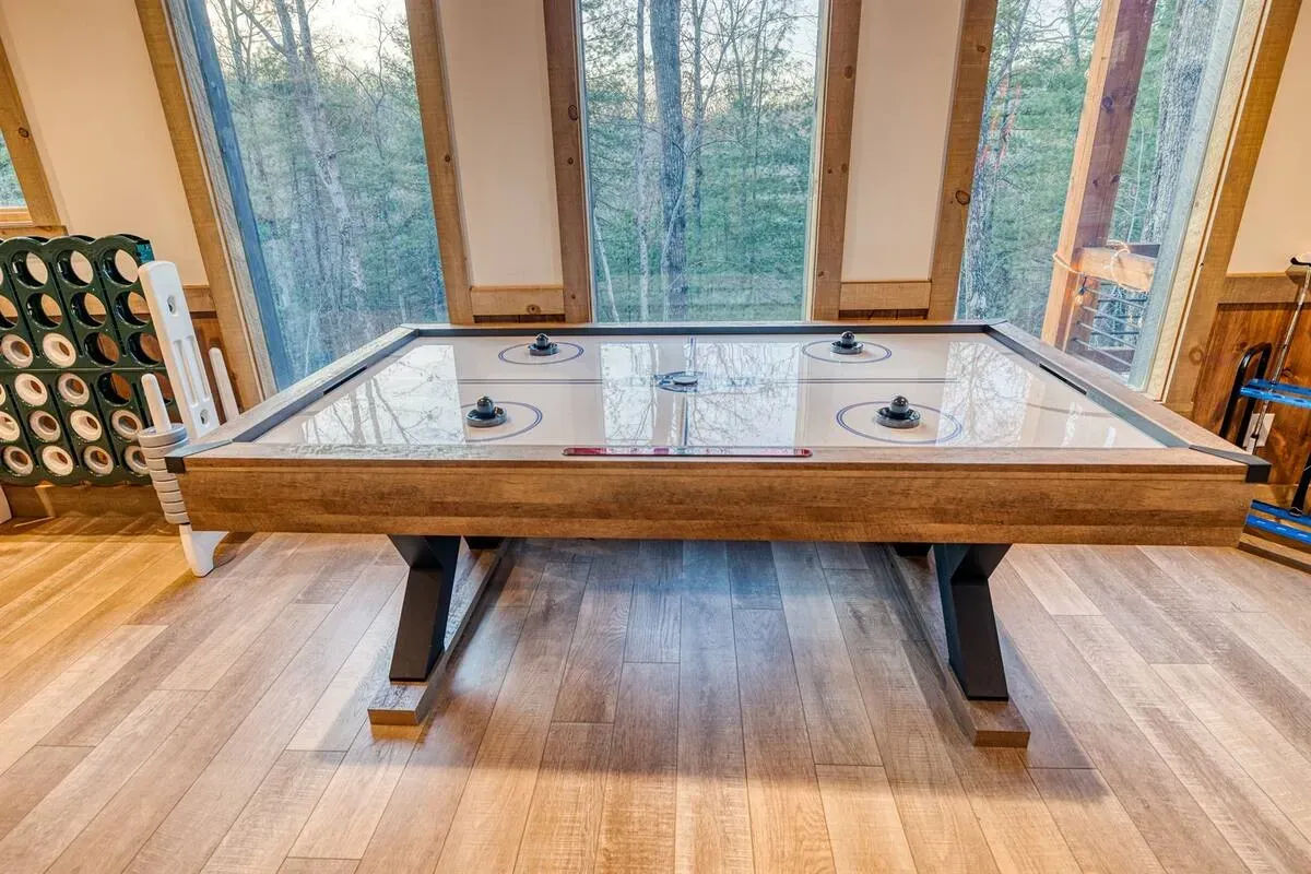 A wooden air hockey table with a glossy white surface sits on a hardwood floor in a room with large windows.