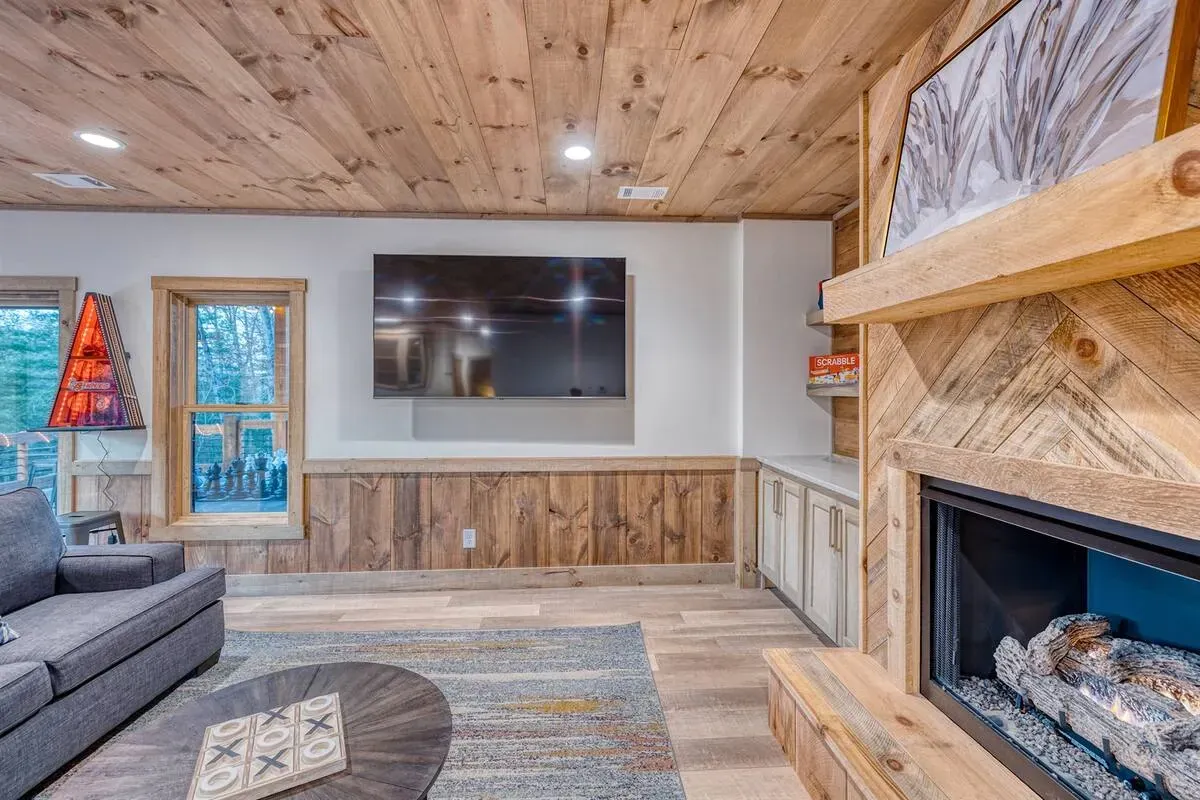 A rustic living room with a wood-paneled ceiling, a wall-mounted TV, a fireplace, and a gray sofa on a patterned rug.