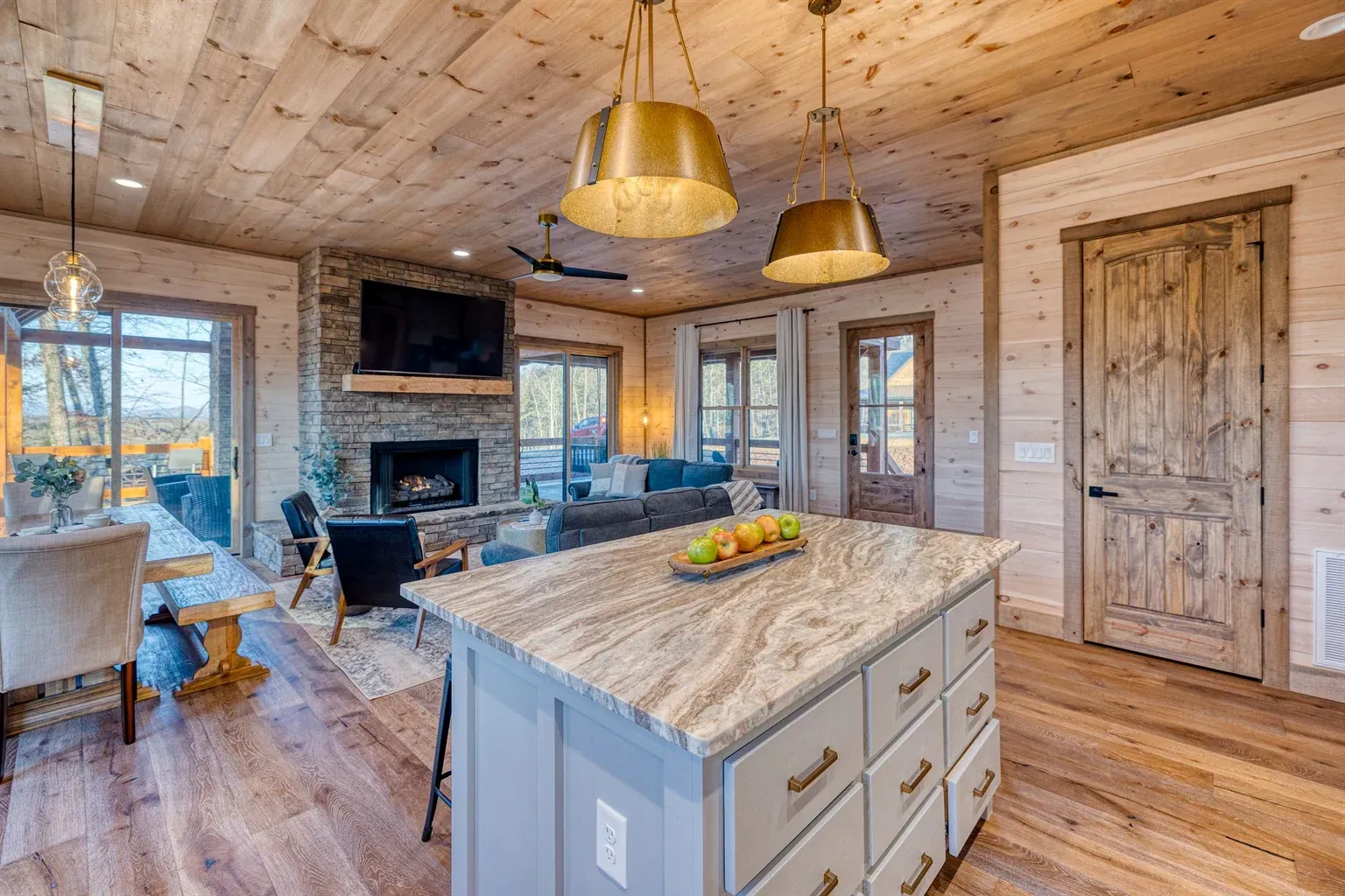 A modern cabin living area featuring a kitchen island with marble countertop, wood paneled walls, and a stone fireplace.