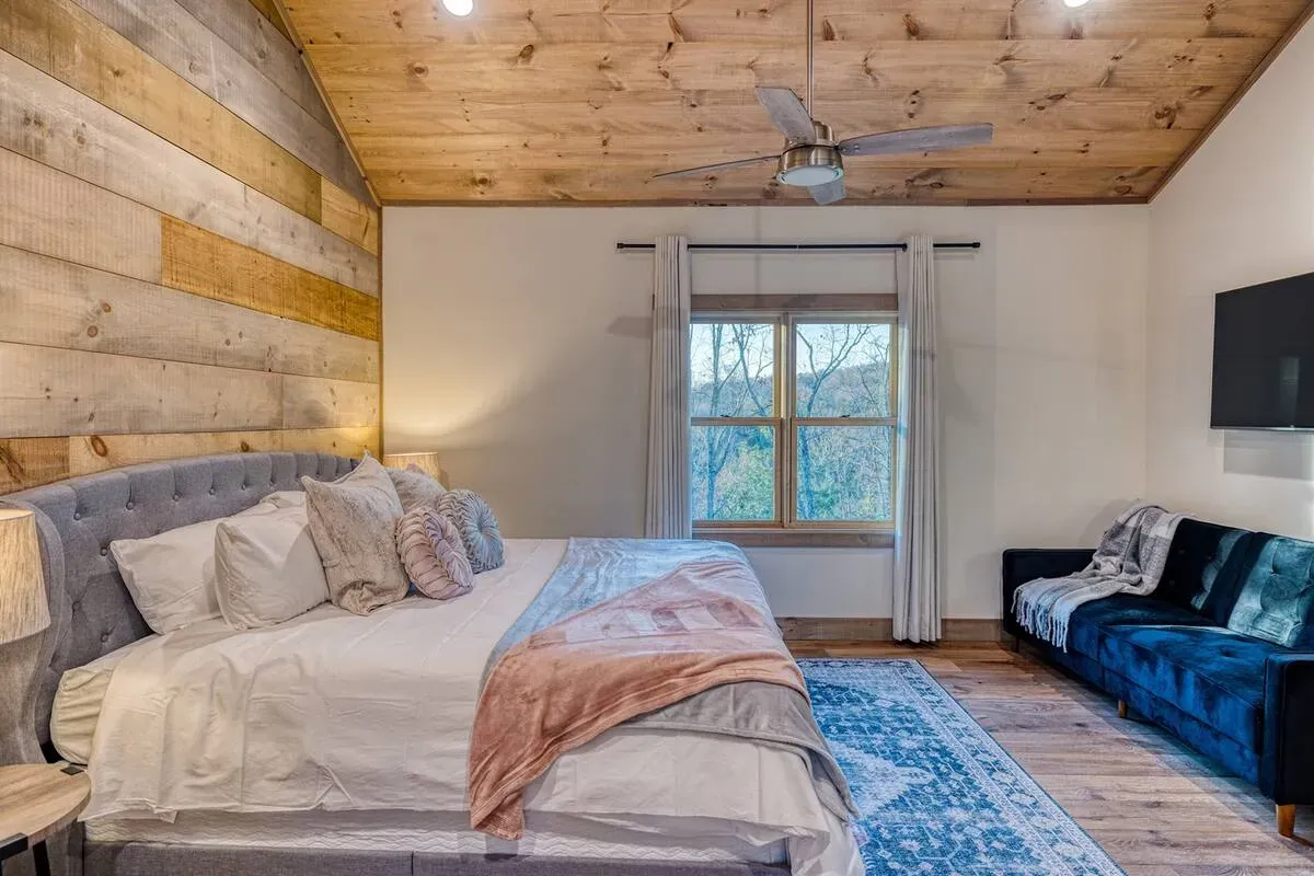 A cozy bedroom with a wooden ceiling, a large tufted bed, a blue velvet sofa, and a window overlooking trees.