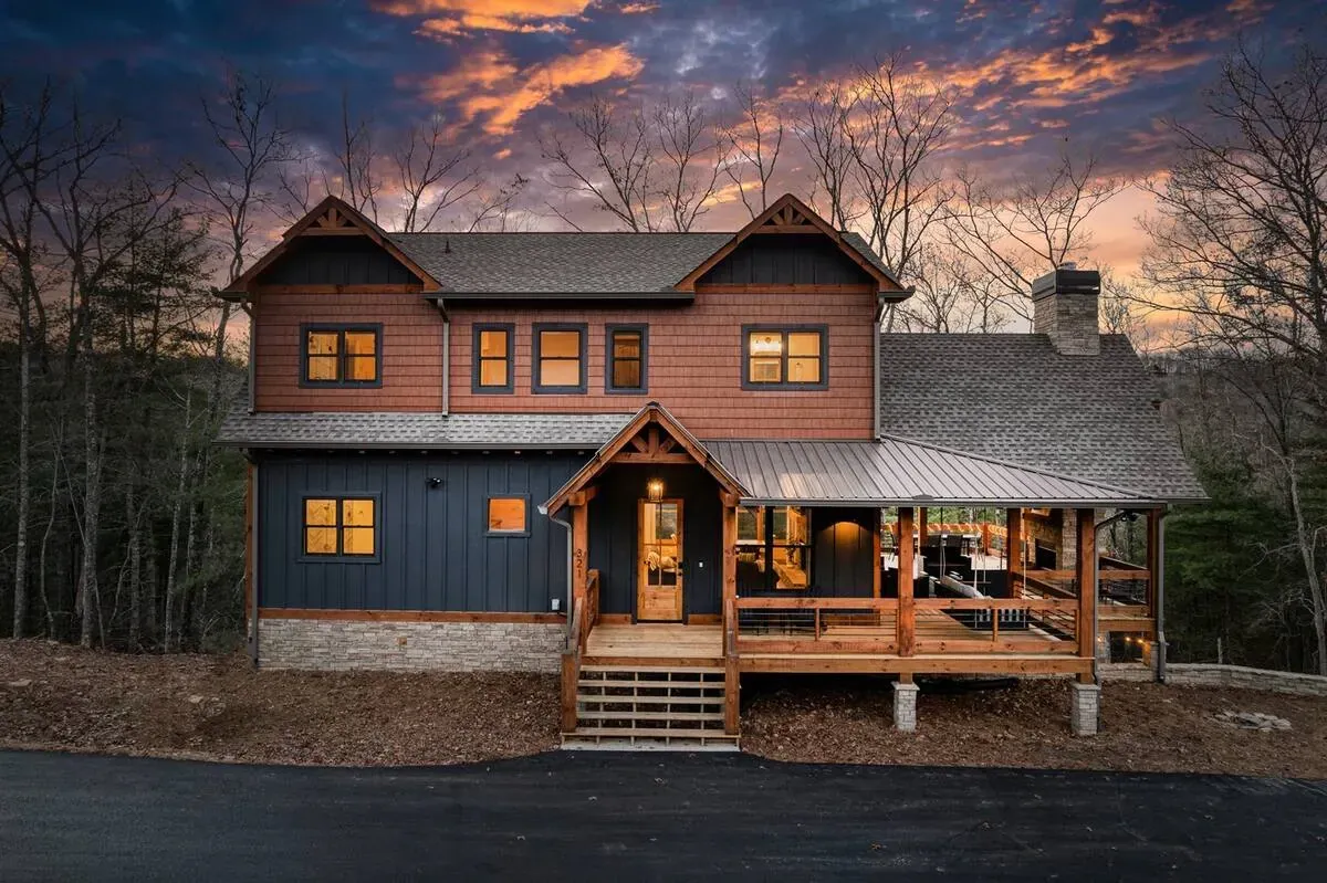 A two-story rustic home with wood and blue siding, a stone foundation, and a spacious covered porch at sunset.