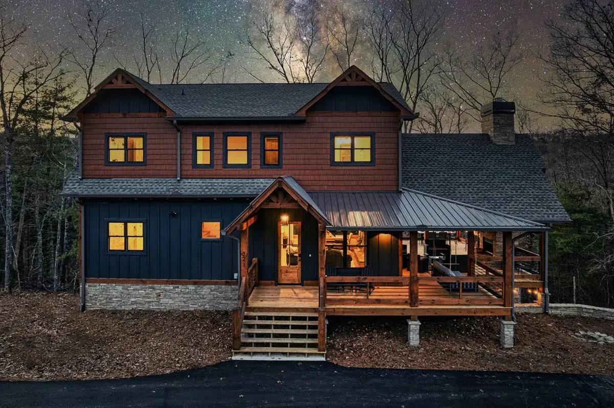 A two-story rustic house with dark blue and wood siding, a stone base, and a lighted porch under a starry night sky.