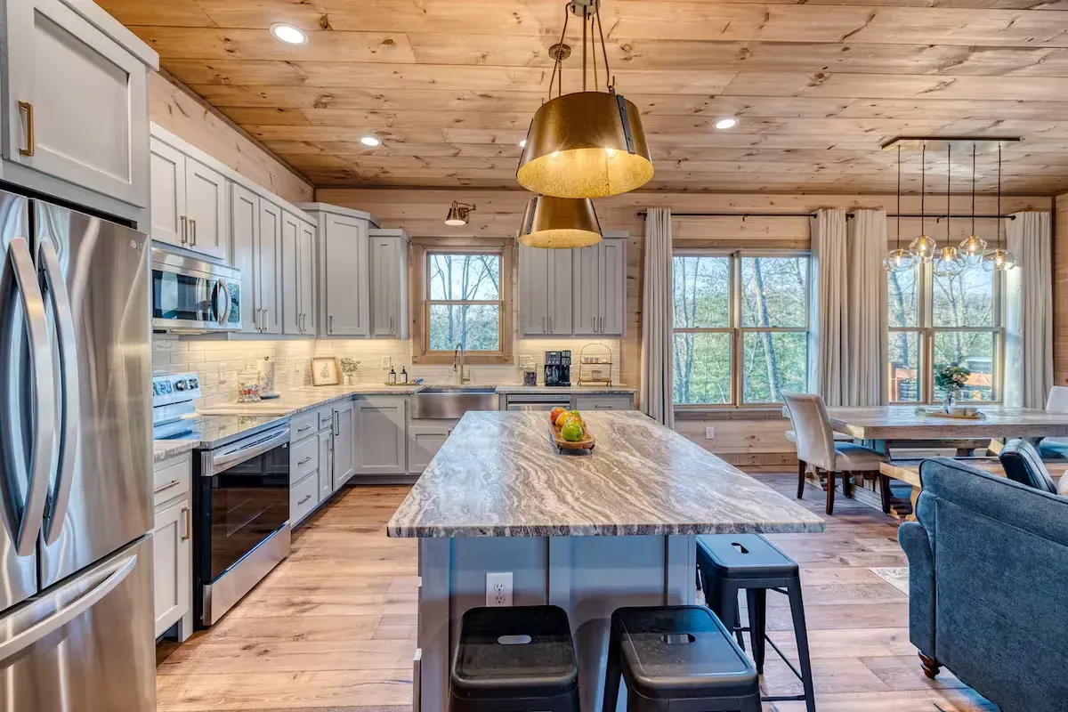 A modern kitchen with white cabinets, a large marble-topped island, wooden ceiling, and pendant lights.