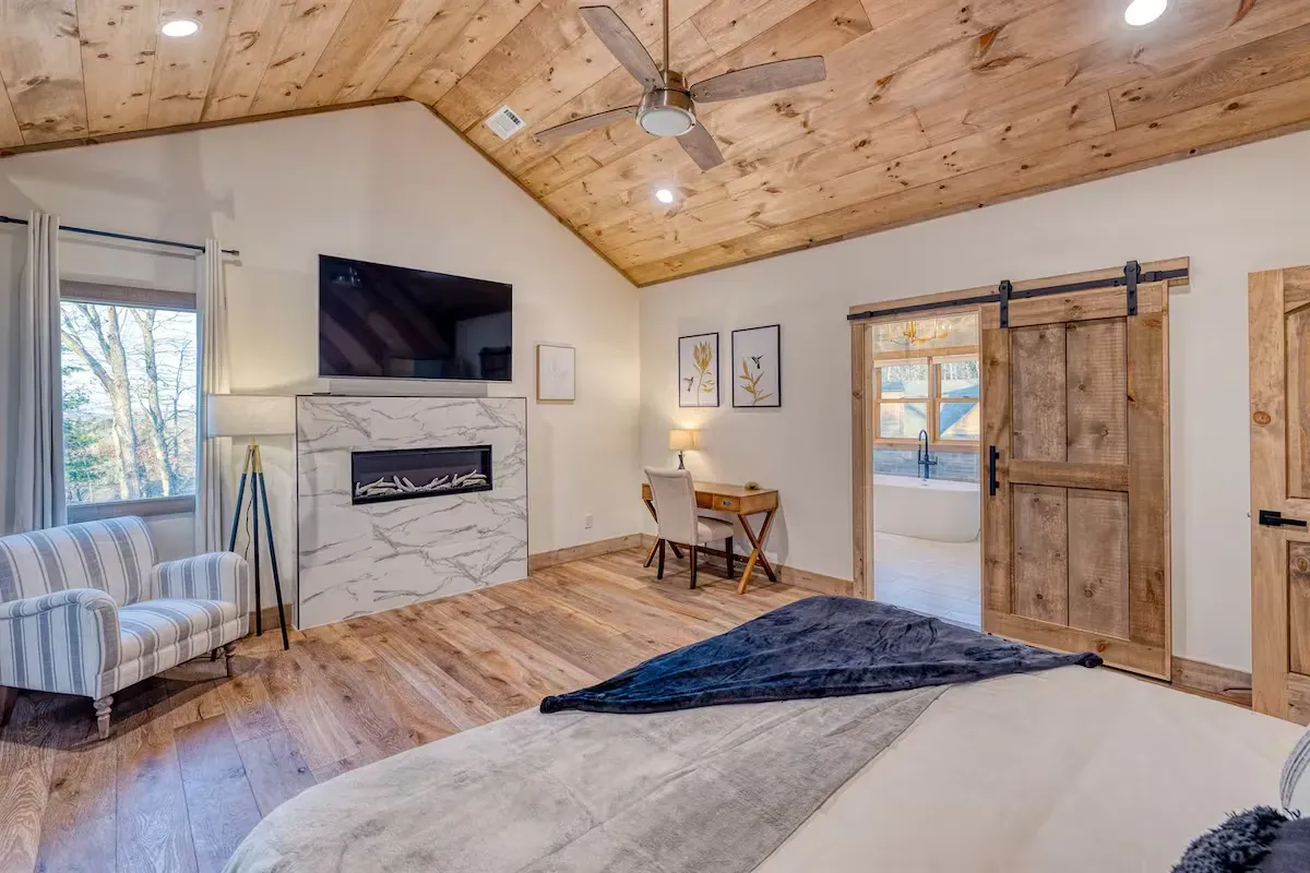 A cozy bedroom with a wood-planked vaulted ceiling, a fireplace with a mounted TV, a sitting chair, and a barn door.