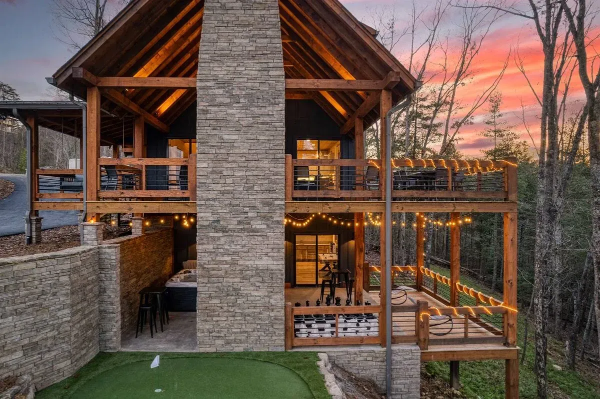 A modern two-story log cabin with a stone chimney, outdoor balconies, string lights, and a putting green at sunset.