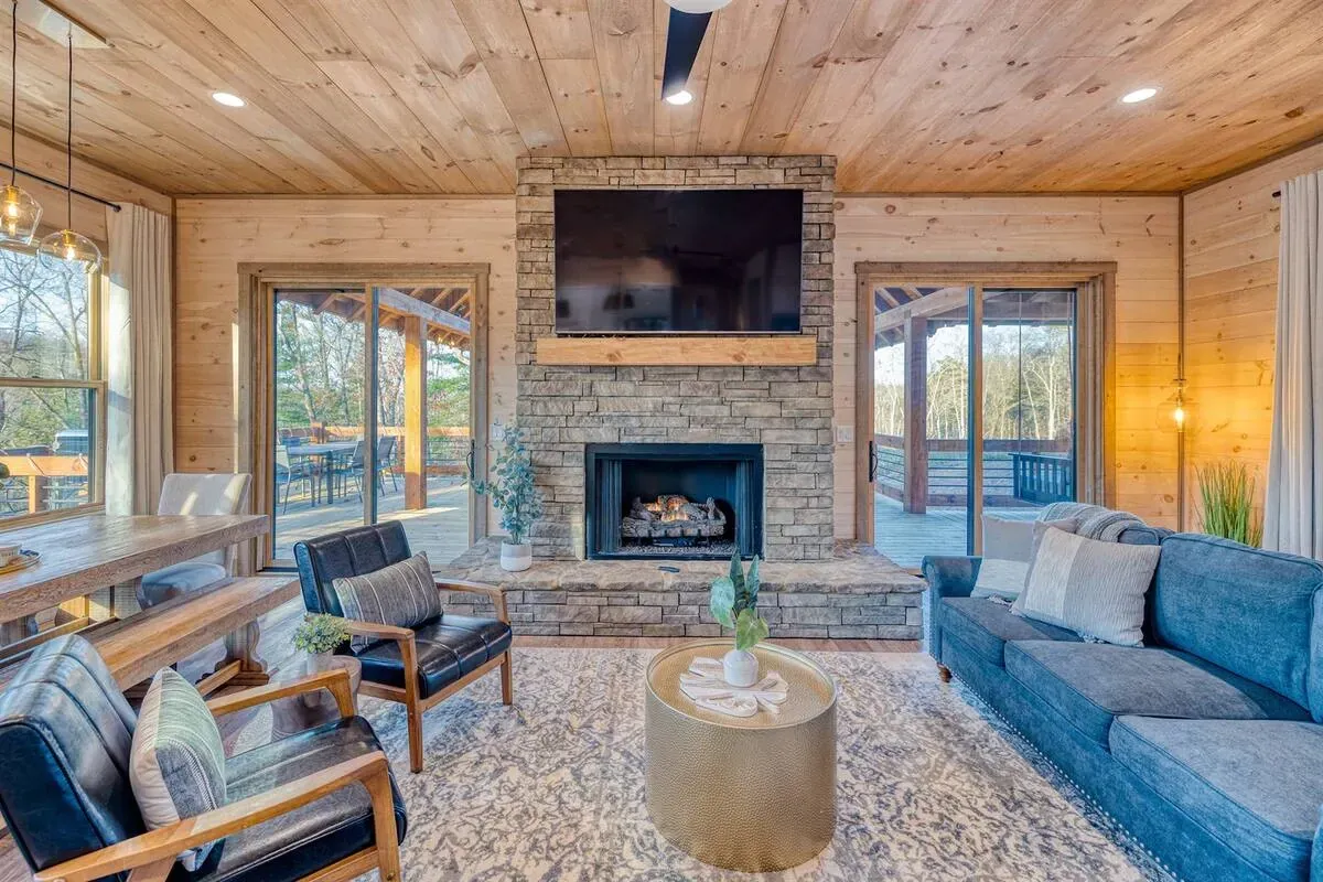A rustic living room with wood paneled walls, a stone fireplace, blue sofa, armchairs, and glass sliding doors.