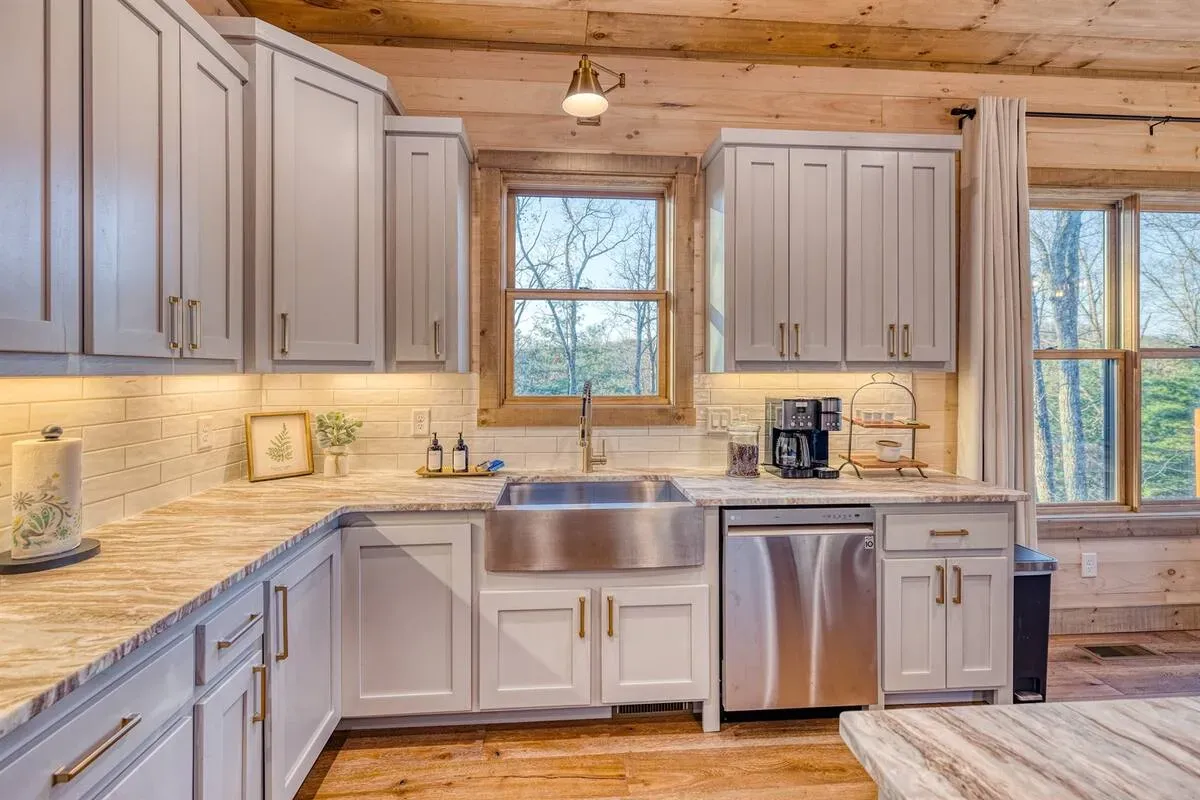A bright, modern kitchen in a log cabin with gray cabinets, a stainless steel farmhouse sink, and natural wood ceilings.