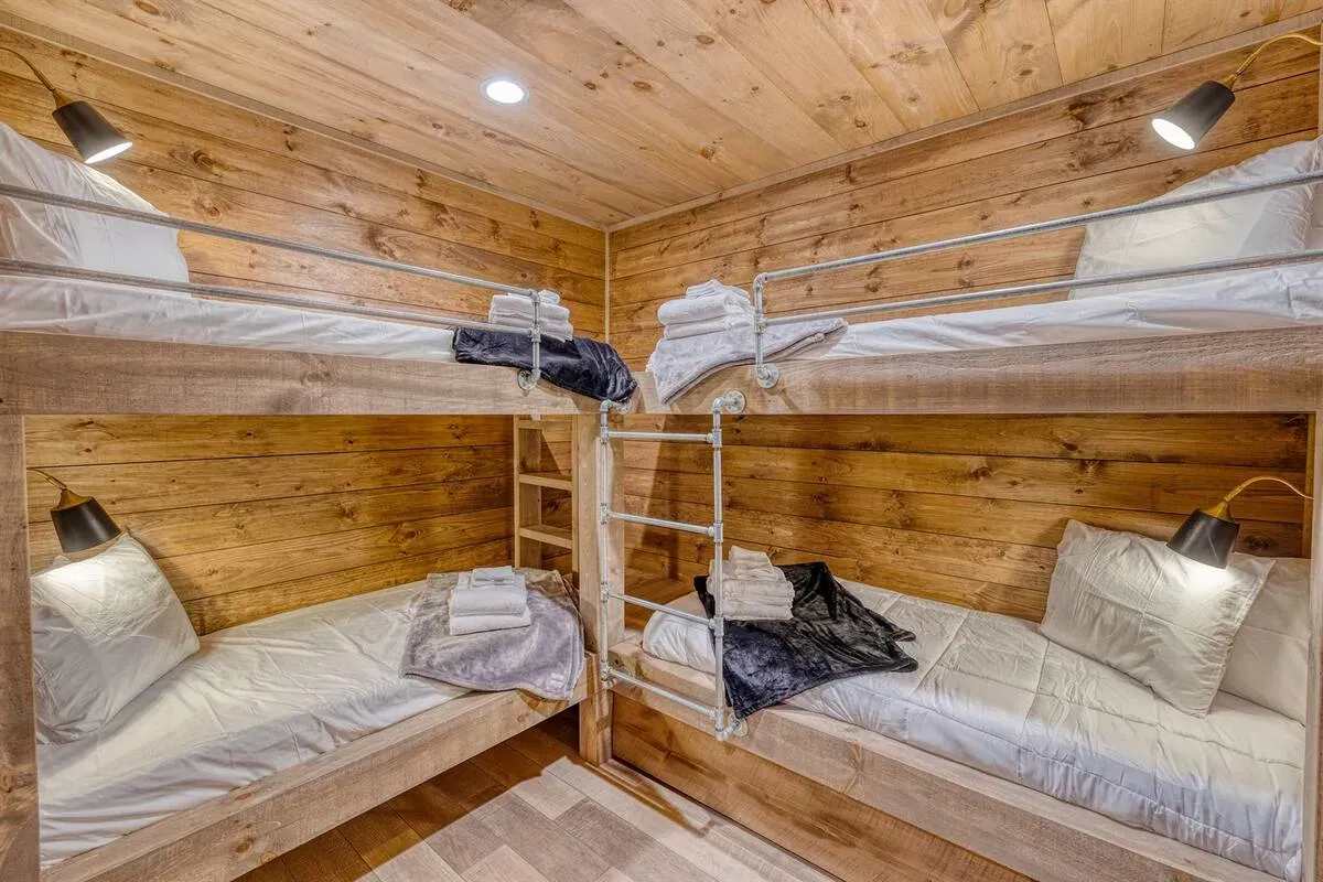 A corner of a room featuring two sets of rustic, light-wood bunk beds with white bedding and small wall-mounted lights.