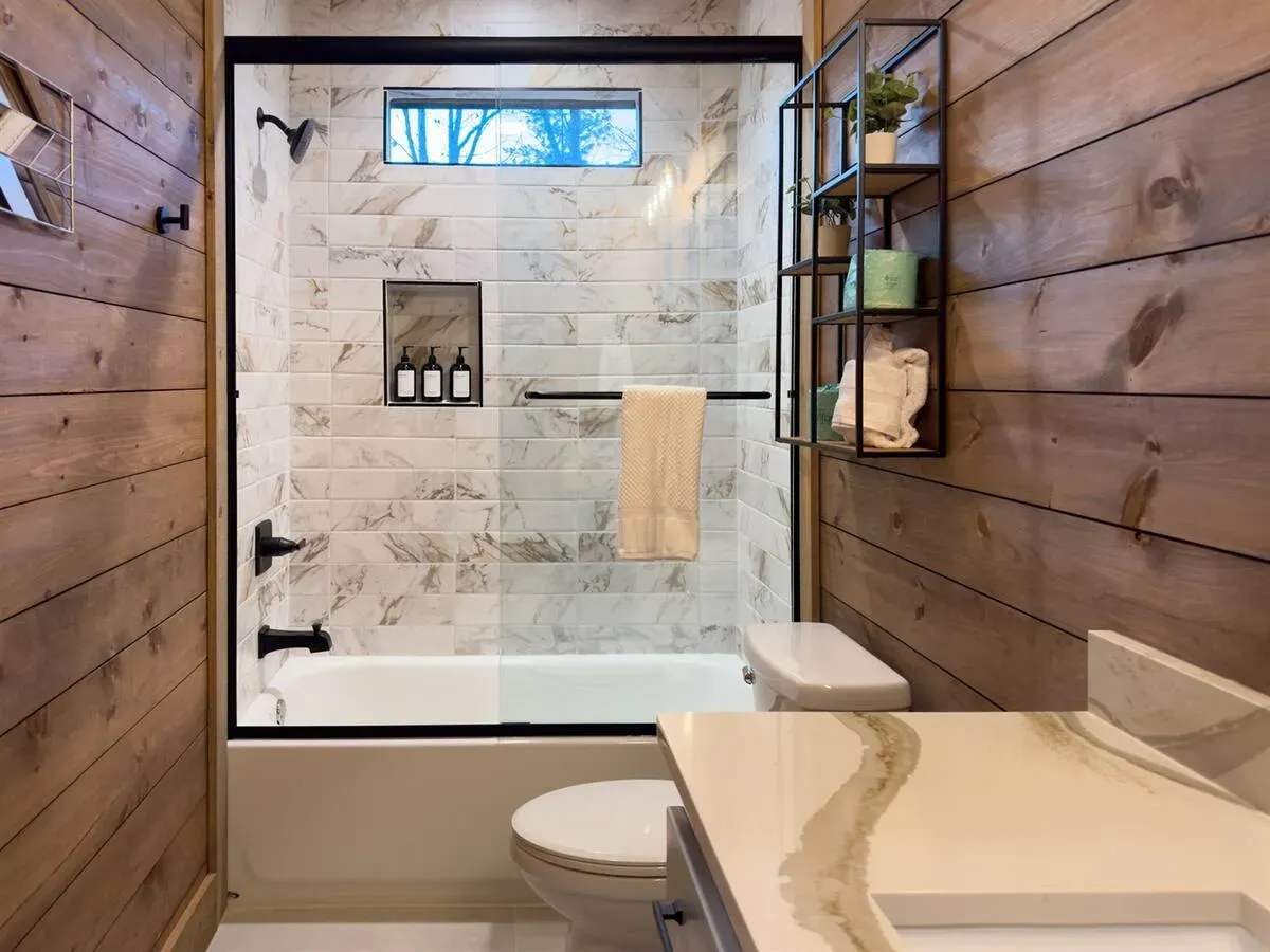 A rustic bathroom featuring wood-paneled walls, a marble-tiled bathtub shower, and a white vanity with a marbled countertop.
