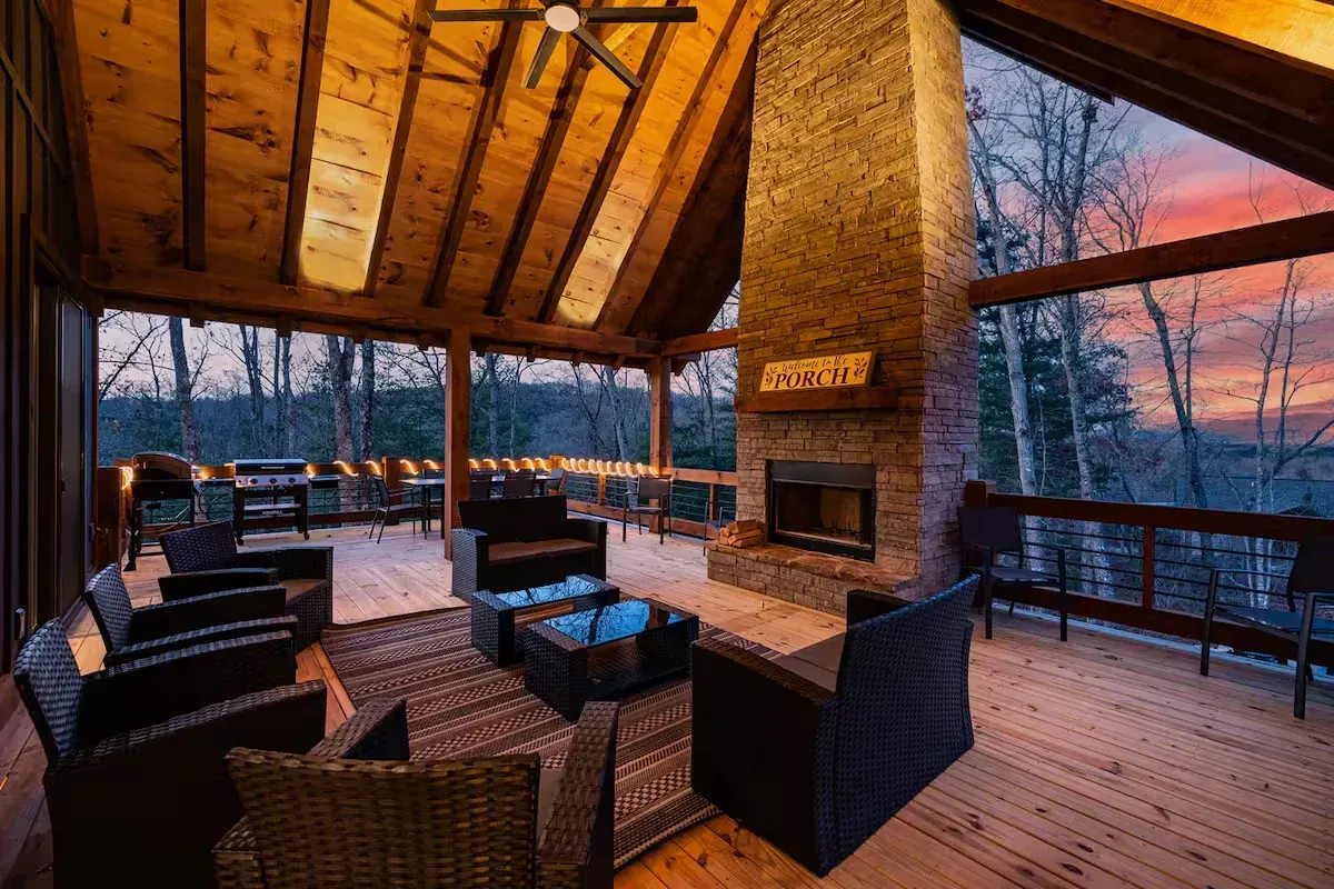 A covered wooden deck features wicker furniture, a stone fireplace, and a panoramic forest view at sunset.