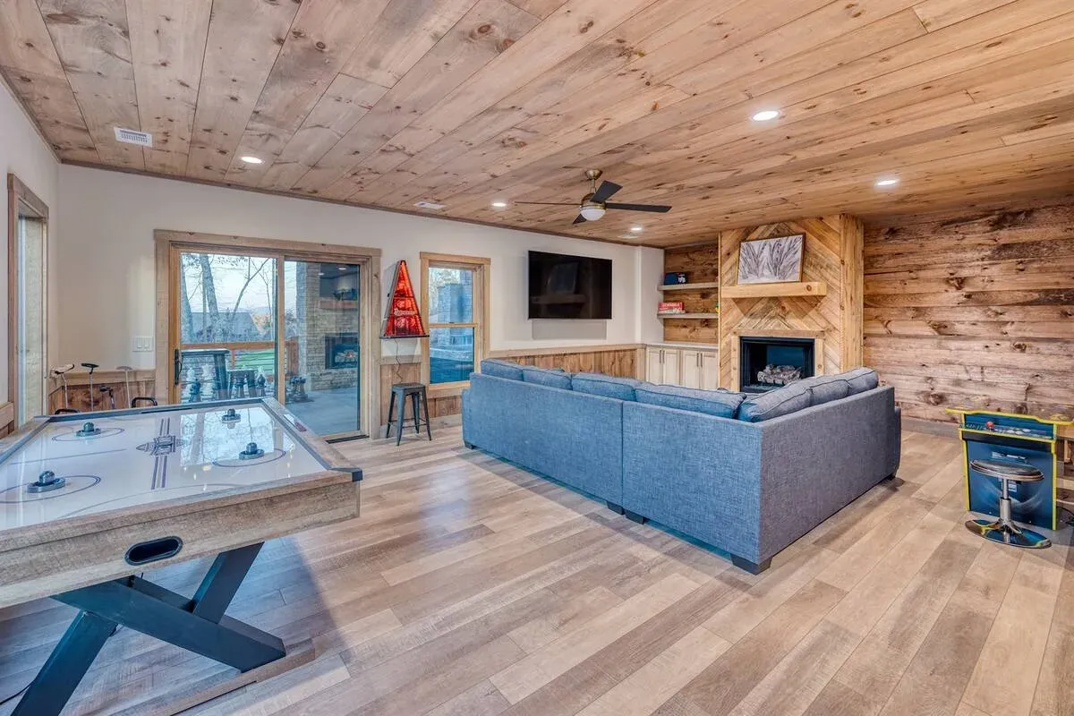 Rustic game room with wood paneling, an air hockey table, a blue sectional sofa facing a fireplace and a mounted TV.