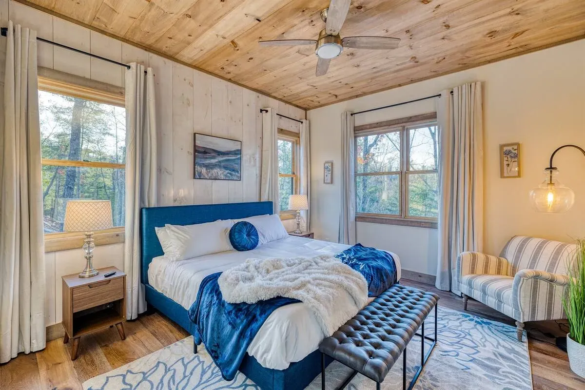 A bright bedroom features a blue-accented bed, a tufted ottoman, wooden ceiling, and windows with white curtains.