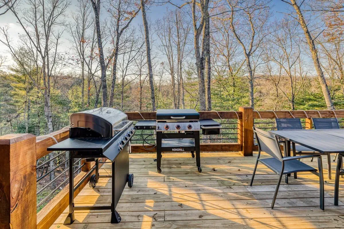 A wooden deck overlooking a forest, featuring a grill and a patio table with chairs under a clear blue sky.