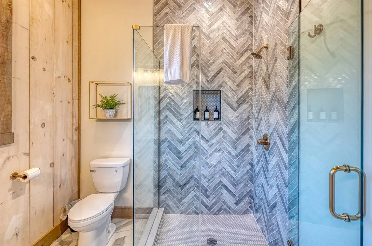 A bathroom featuring a glass-enclosed shower with grey herringbone tile, a white toilet, and light-toned wood paneled walls.