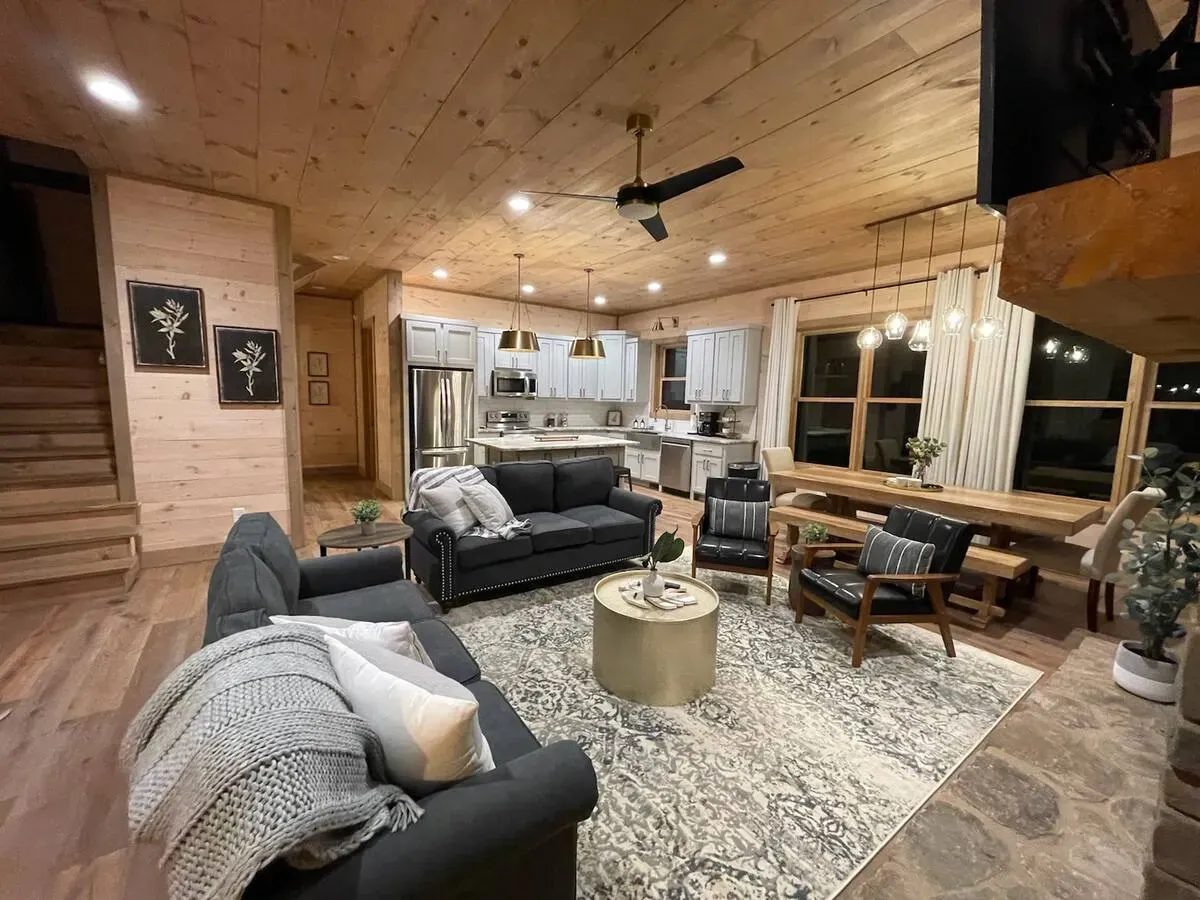 A rustic, open-concept cabin living room with wooden walls, dark sofas, a round gold coffee table, and an adjacent kitchen.