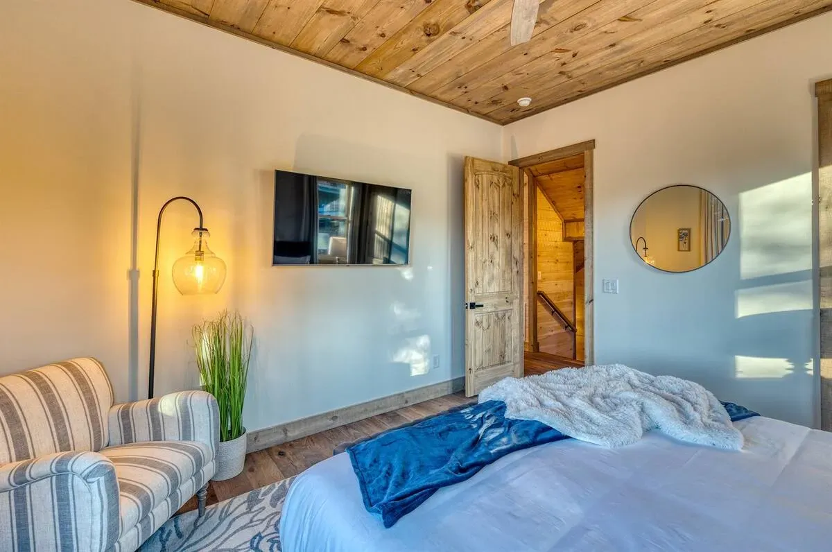A bedroom with wood-paneled ceiling, a bed with blue bedding, a striped armchair, floor lamp, and wall mirror.