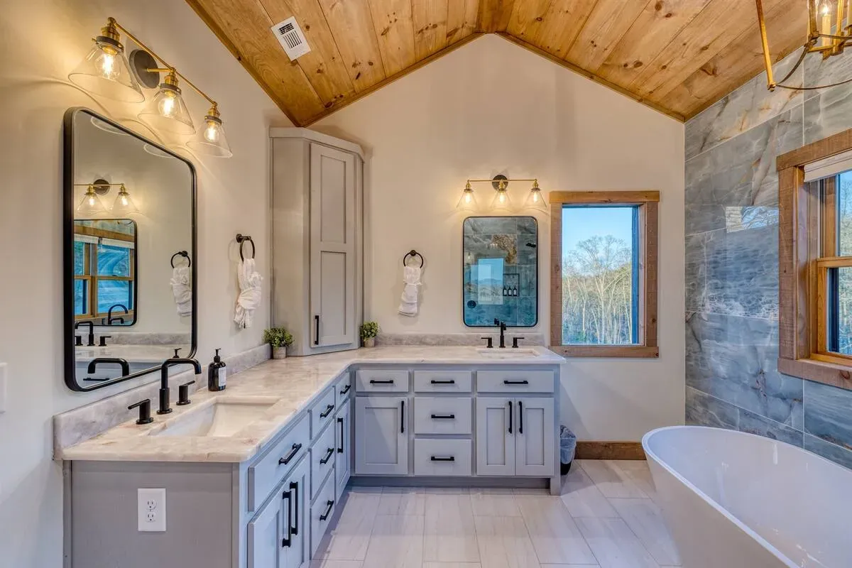A bright, modern bathroom featuring grey vanities, a white freestanding tub, dual mirrors, and a vaulted wood ceiling.