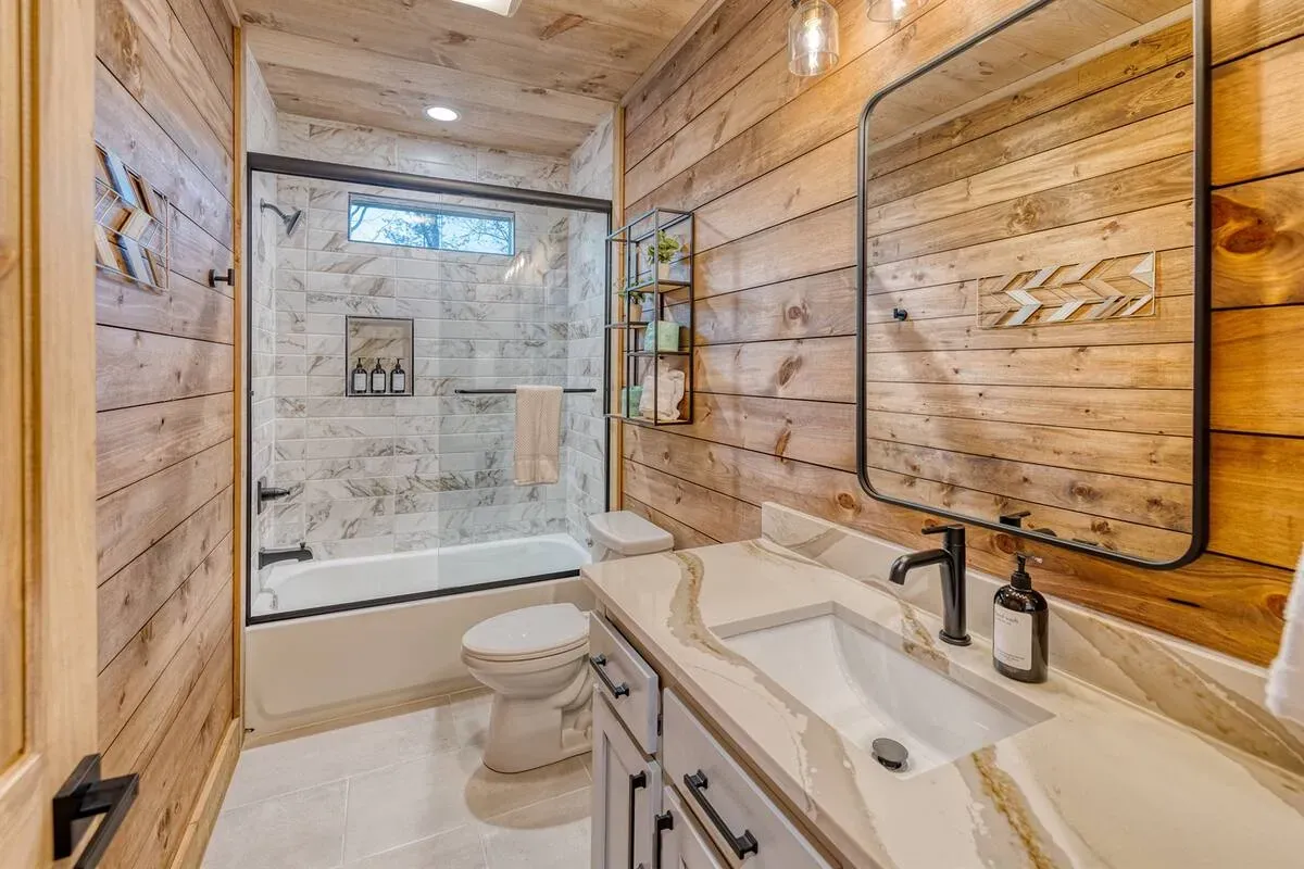 Modern rustic bathroom with wood plank walls, white marble-style shower, vanity with stone countertop, and black hardware.