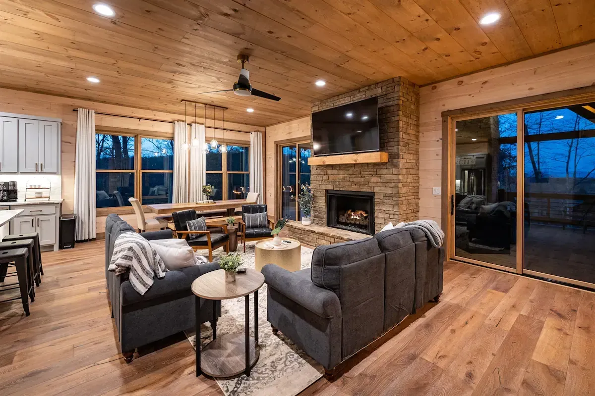 A cozy living room with grey sofas, a stone fireplace with a TV, wood-paneled ceiling, and large windows at twilight.