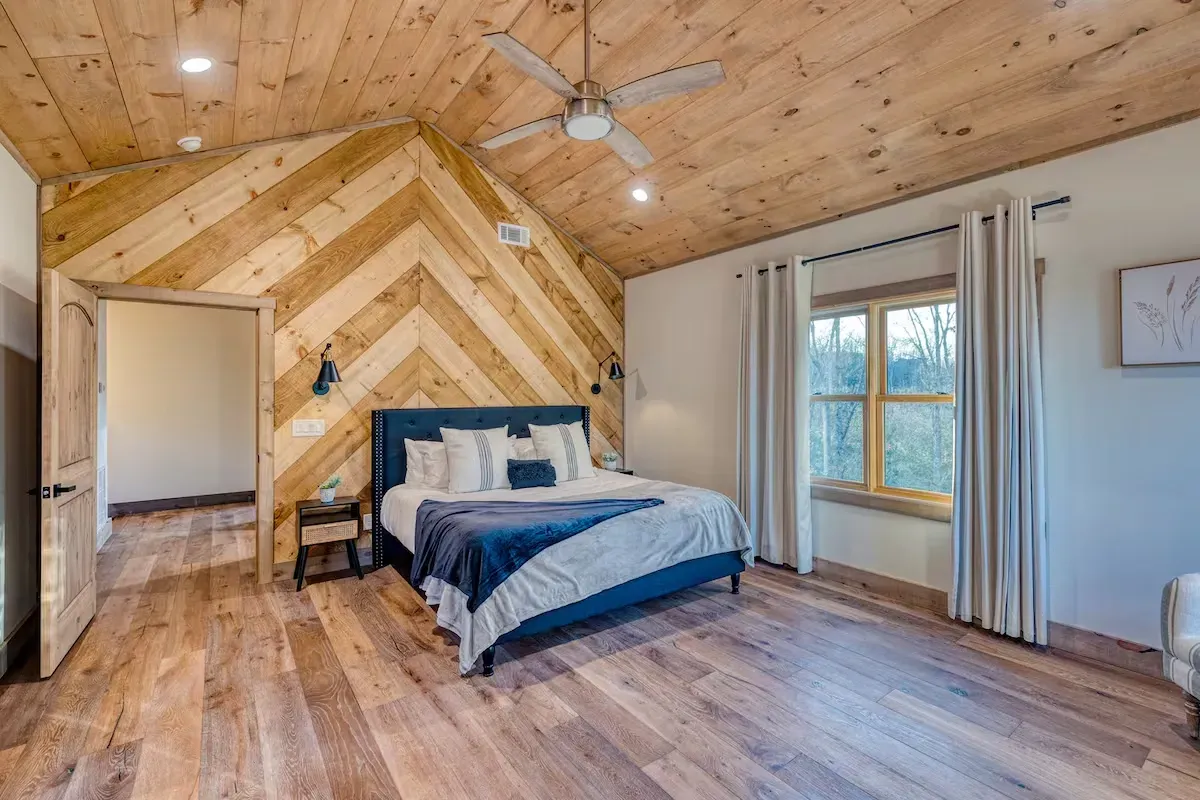 A bedroom featuring a blue bed, light wood floors, a chevron-patterned wood accent wall, and a large window with curtains.